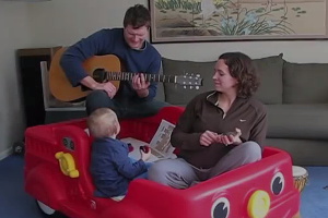 A man playing guitar while two women and a baby sit in a toy car.